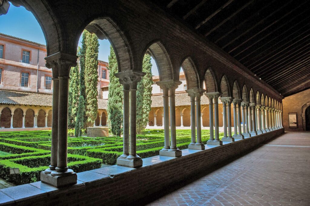 Serene view of the cloister at Couvent des Jacobins in Toulouse, featuring a long corridor with elegant stone arches and columns. The peaceful courtyard is adorned with symmetrical green hedges and tall cypress trees, creating a harmonious blend of architecture and nature.