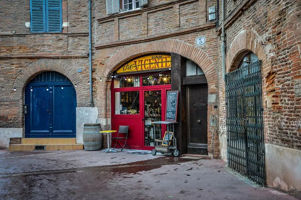 Charming entrance of Chez Vicente, a traditional bodega in Toulouse, featuring a bright red storefront with a yellow sign. The cozy establishment is nestled between brick arches, with a small outdoor seating area including a table and chairs, and a chalkboard menu displayed outside