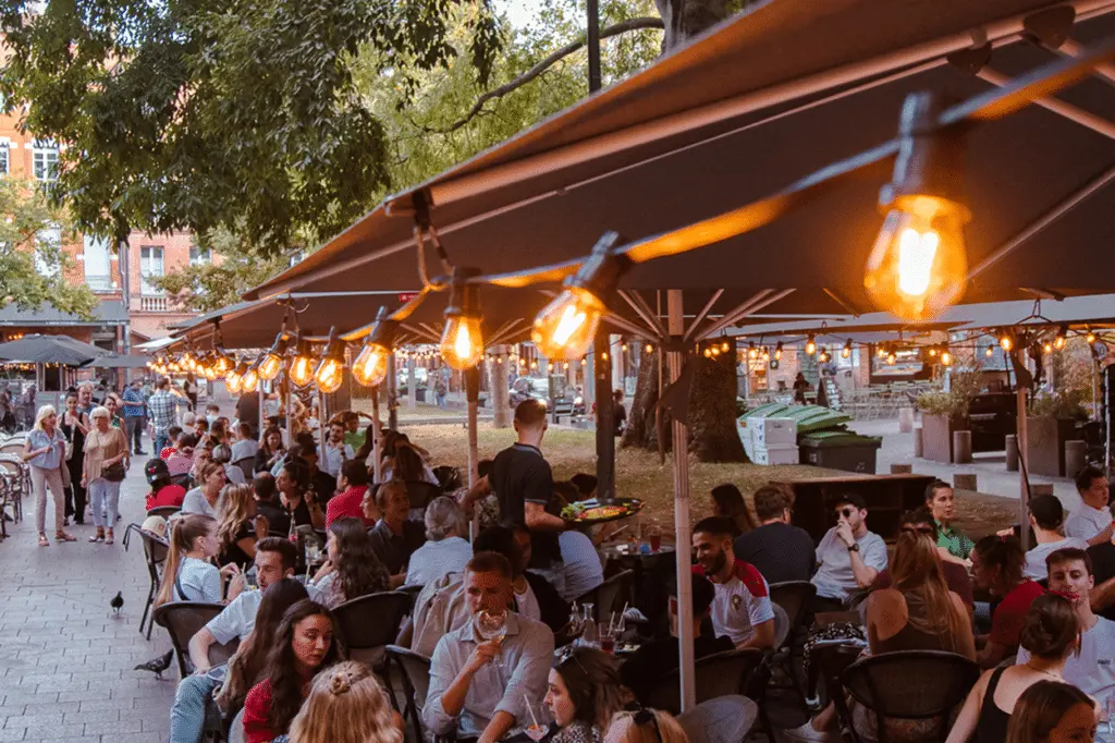 Lively outdoor terrace of a café or restaurant in Toulouse, filled with people enjoying drinks and conversations under string lights. The space features large umbrellas, wooden tables, and chairs, creating a vibrant and convivial atmosphere in an urban setting with trees and buildings in the background.