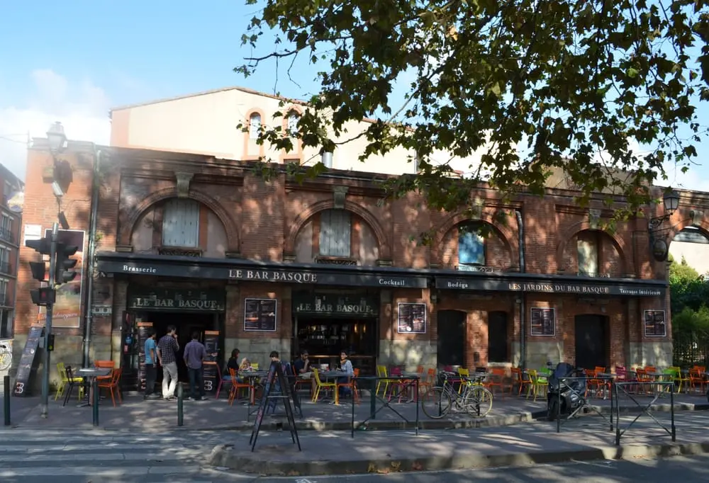 Exterior view of Le Bar Basque in Toulouse, a charming establishment with a traditional brick facade and arched windows. The terrace is set up with colorful chairs and tables, where patrons are seen enjoying drinks and conversation under the shade of trees on a sunny day.