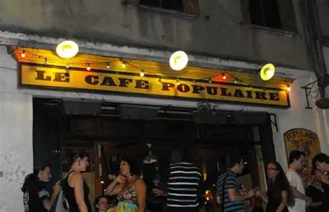Evening scene at the entrance of Le Café Populaire, a lively café illuminated by warm yellow lights. A group of people is gathered inside and outside the entrance, socializing and enjoying the vibrant atmosphere of this popular nightspot.