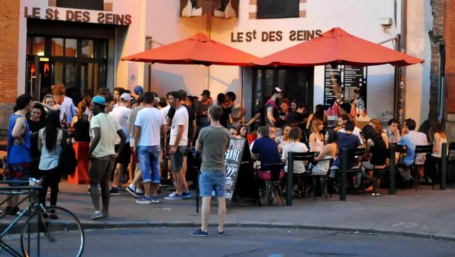 Busy evening at Le Saint des Seins bar in Toulouse, with a crowd of people socializing both at outdoor tables under red umbrellas and standing around the entrance. The lively atmosphere captures the essence of a popular nightlife spot, with patrons enjoying drinks and conversations.