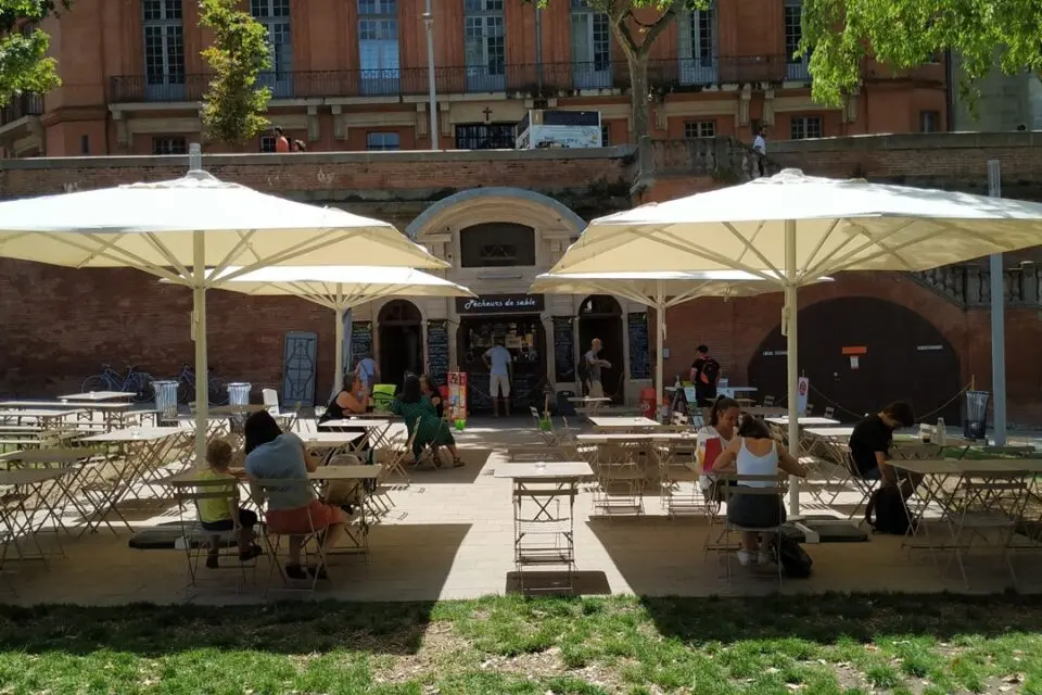 Sunny outdoor terrace of the restaurant Les Pêcheurs de Sable in Toulouse, featuring large white umbrellas shading wooden tables and chairs. Patrons are seen enjoying their meals and the pleasant atmosphere, with a view of the historic brick building and lush greenery in the background.
