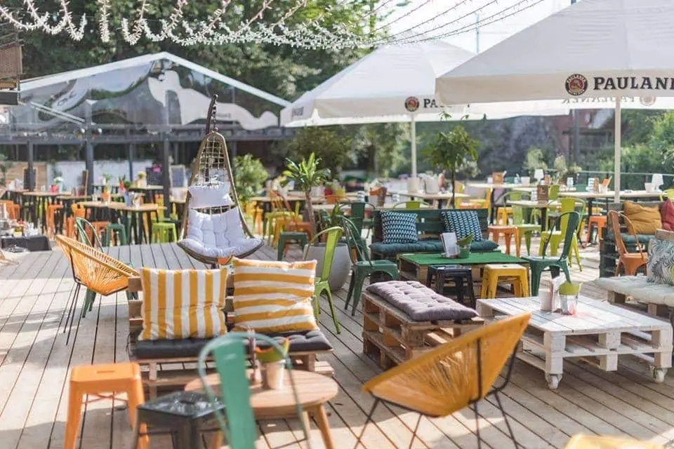 Colorful and inviting rooftop terrace in Toulouse, featuring a variety of seating options including striped cushions, hanging chairs, wooden pallet sofas, and metal stools. The space is decorated with green plants, string lights, and large umbrellas branded with 'Paulaner', creating a cozy and vibrant atmosphere for enjoying Toulouse by night.
