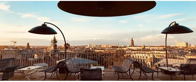 Stunning rooftop terrace in Toulouse during sunset, featuring modern black lounge chairs, small tables, and stylish black lamps. The panoramic view showcases the city's skyline, including historic buildings and church towers, under a clear sky with warm golden light.