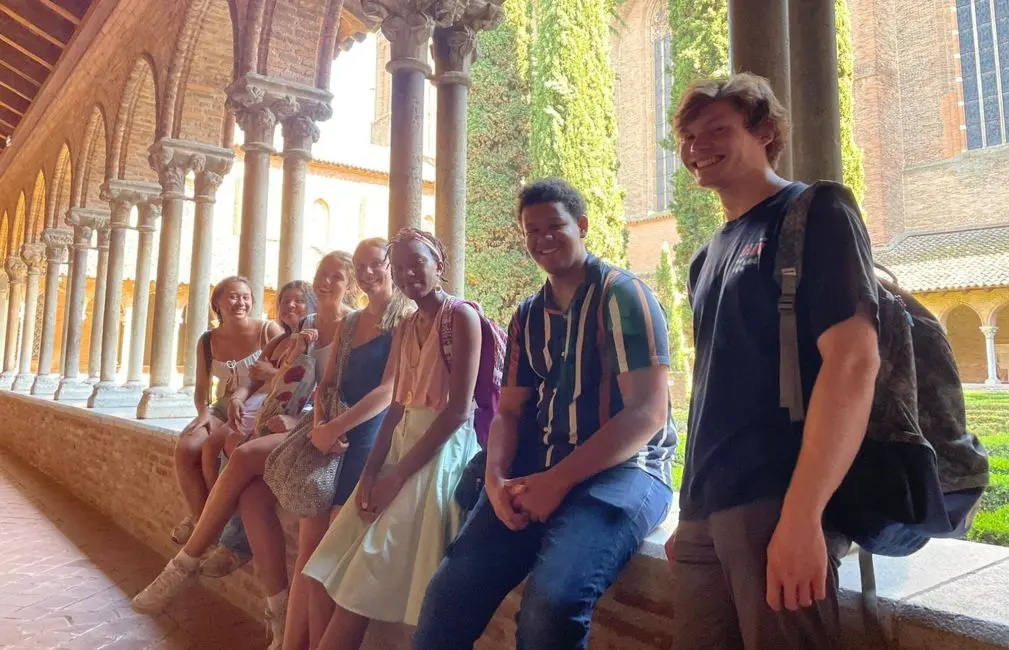 Group of smiling students posing together in the cloister of a historic building in Toulouse. The scene features a row of ornate columns and arches in the background, with lush green ivy climbing the walls, creating a picturesque and serene atmosphere.