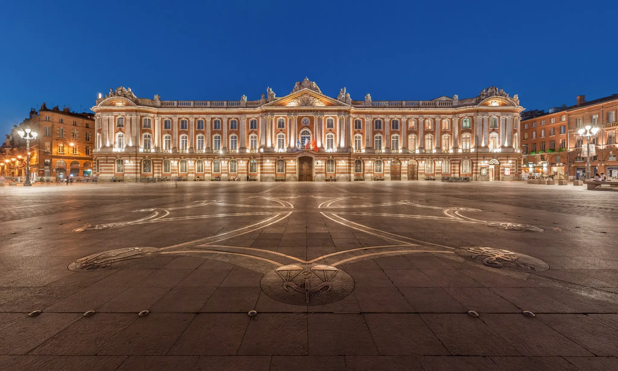 Nighttime view of Place du Capitole in Toulouse, France, showcasing the grand, illuminated Capitole building with its neoclassical architecture and warm brick facade. The spacious, paved square features the iconic Occitanie cross and zodiac symbols, with street lamps casting a soft glow over the serene, almost empty plaza.