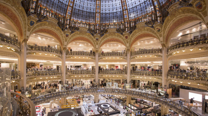 Grand interior view of a luxurious multi-level department store, featuring an impressive glass-domed ceiling that allows natural light to flood the space. The store is adorned with ornate golden arches, intricate railings, and multiple floors filled with a variety of high-end boutiques and displays. Shoppers can be seen browsing the aisles, adding to the bustling and opulent atmosphere of this historic shopping destination.