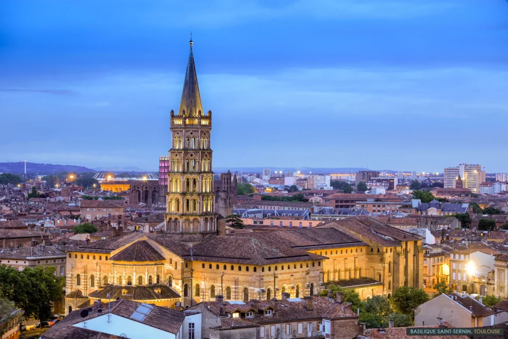 Evening view of Toulouse skyline featuring the illuminated Basilica of Saint-Sernin, with its iconic bell tower and Romanesque architecture. The cityscape includes terracotta rooftops, historic buildings, and a softly lit urban panorama at dusk, showcasing the charm of the Pink City.
