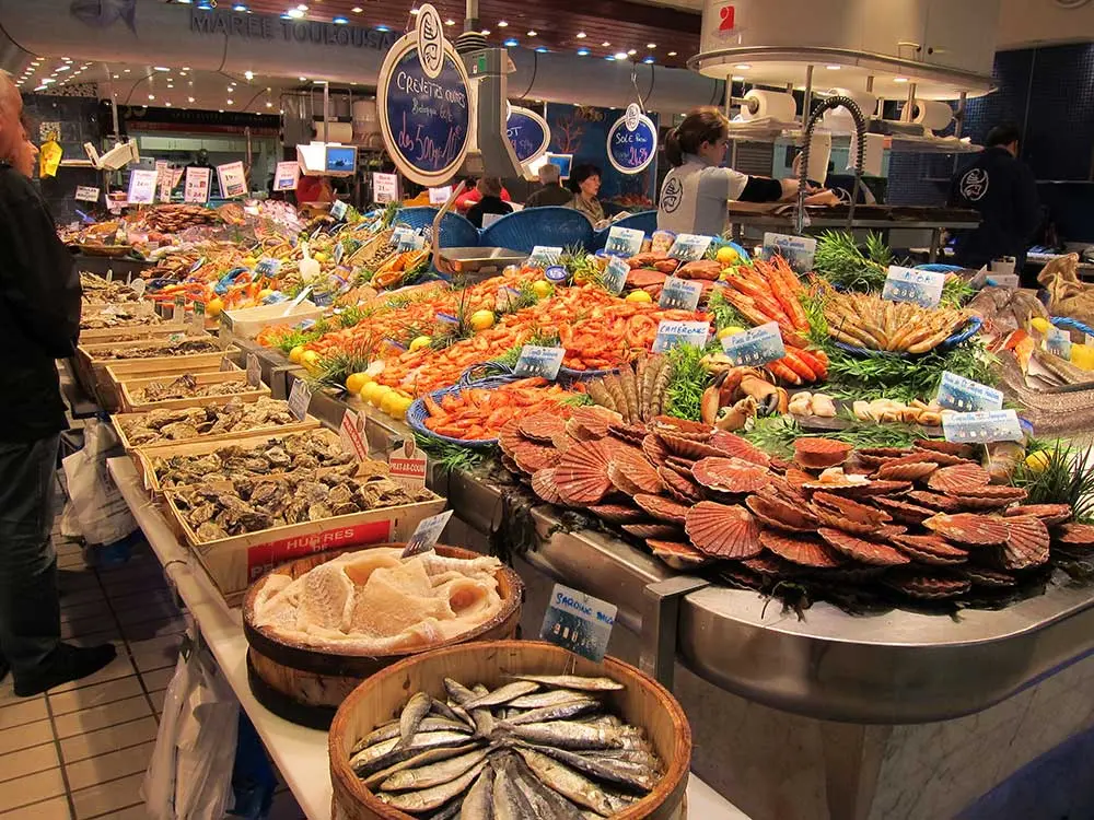 Vibrant seafood display at a market in Toulouse, featuring a wide variety of fresh offerings such as oysters, scallops, shrimp, crab, clams, and sardines. The colorful arrangement includes ice beds, wooden baskets, and handwritten price signs, with vendors and customers interacting in the background, creating a lively and appetizing atmosphere.