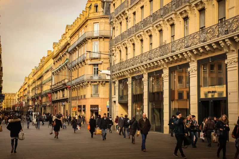 Busy street scene in Toulouse during the day, featuring a row of elegant, historic buildings with ornate facades and wrought-iron balconies. Pedestrians are seen walking, shopping, and interacting in front of various shops and boutiques, creating a lively and vibrant urban atmosphere. The warm lighting of the late afternoon enhances the charm of the architecture and the bustling activity.