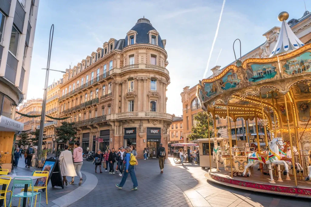 Lively street scene in Toulouse during the day, featuring a beautifully ornate vintage carousel with horses and decorative lights on the right. In the background, a grand historic building with intricate architectural details and wrought-iron balconies houses modern shops like Snipes. People are seen walking, chatting, and enjoying the outdoor cafés, adding to the vibrant and festive atmosphere of the city center.