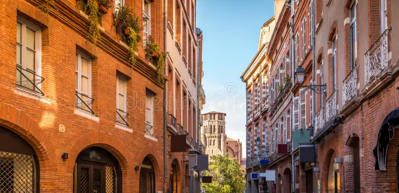 Charming narrow street in Toulouse, lined with traditional red-brick buildings featuring wooden shutters and wrought-iron balconies. The street leads towards a historic bell tower in the distance, under a clear blue sky. The warm sunlight enhances the rustic beauty of the architecture, creating a picturesque and inviting urban scene.