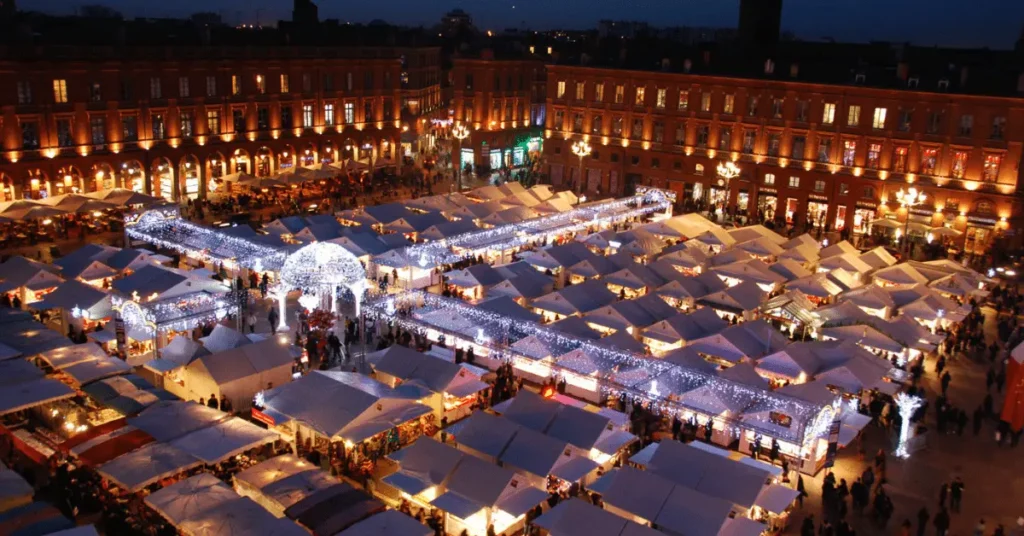 Aerial nighttime view of a bustling Christmas market in Toulouse, featuring rows of illuminated white stalls adorned with twinkling fairy lights. The market is set against the backdrop of historic red-brick buildings, creating a festive and magical atmosphere. Visitors are seen strolling among the stalls, enjoying the holiday spirit under the warm glow of the decorations.